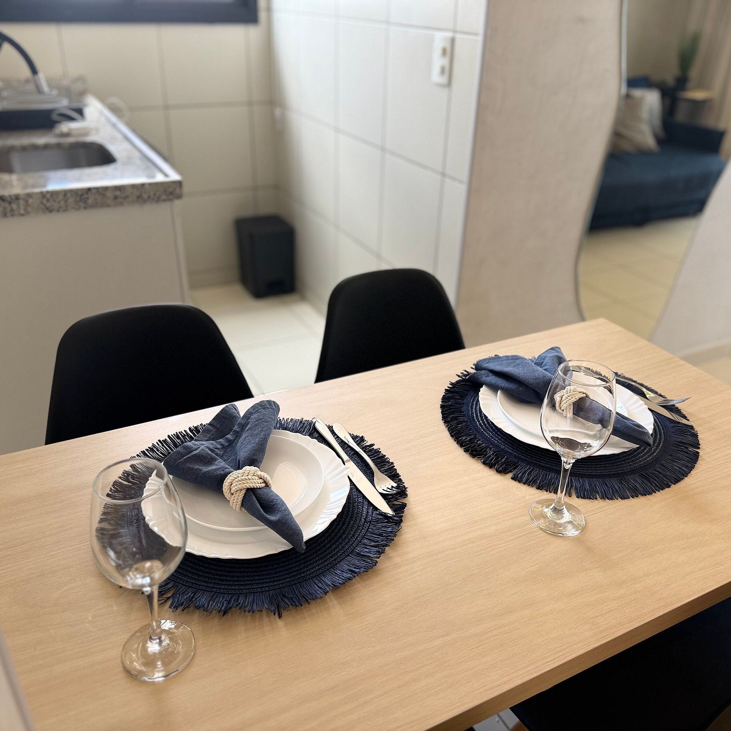 Una mesa de comedor dispuesta con platos, cubiertos y copas de vino, con un altavoz y una pared de azulejos al fondo.