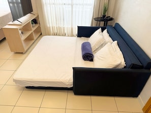 A neatly made bed with white linens and a rolled purple towel, a dark blue sofa with white cushions, a small wooden side table with a potted plant, and a television on a wooden stand. - Loft em Ponta Negra com vista para o mar (Natal)