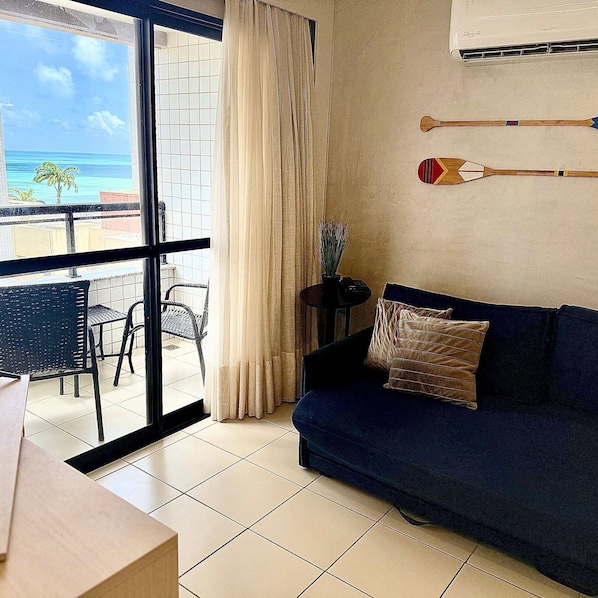 A room with a dark blue sofa, a small table, and a chair. There is a window with a view of the ocean and palm trees. - Loft em Ponta Negra com vista para o mar (Natal)