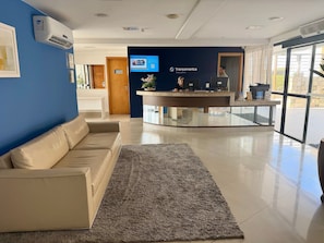 A modern reception area with a blue wall, a beige sofa, a glass counter, and a digital screen displaying a welcome message. - Loft em Ponta Negra com vista para o mar (Natal)