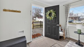 Una habitación con una puerta negra, un banco de madera, una mesita con una lámpara y vistas al exterior.