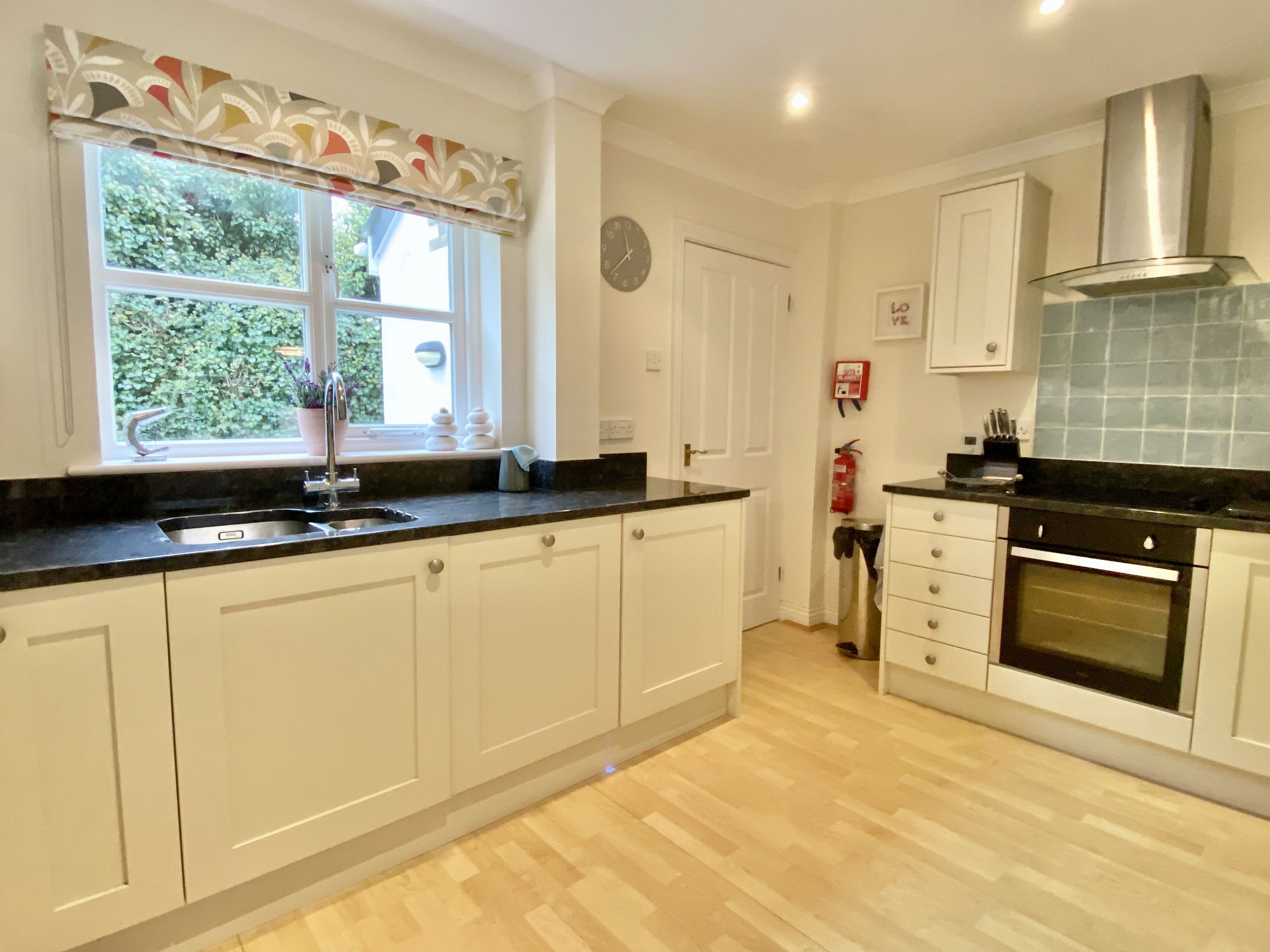 A kitchen with white cabinets, a black countertop, a stainless steel range hood, a window with a decorative valance, and a fire extinguisher mounted on the wall.