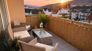 Un balcon agrémenté de meubles en osier, d’une table en verre et offrant une vue sur la ville au coucher du soleil.