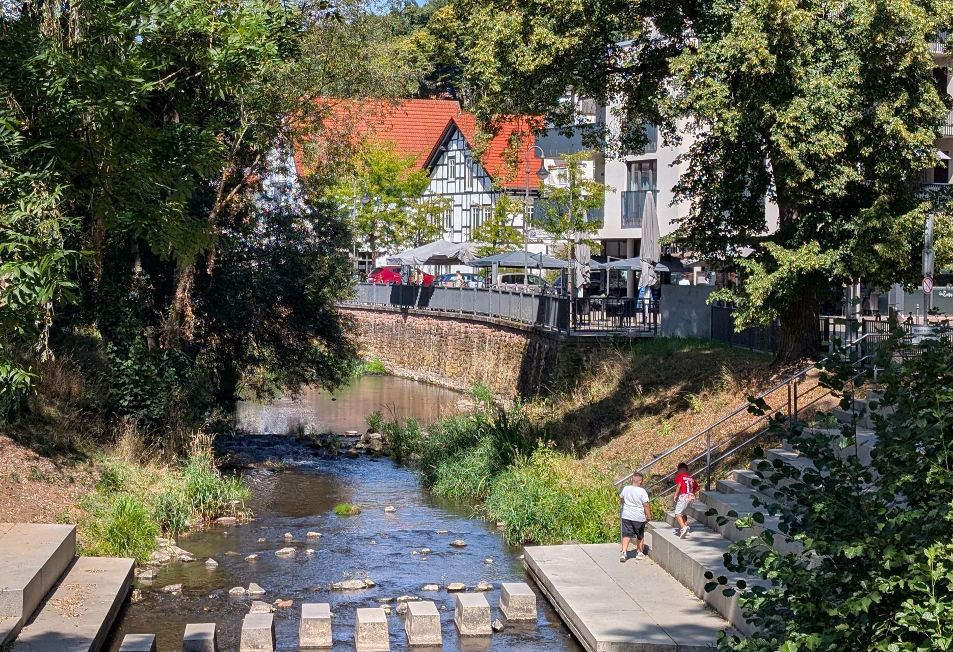 A river with a stone bridge, a pedestrian walkway, and buildings in the background.