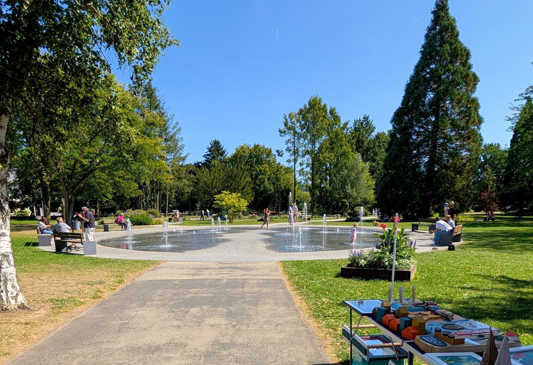 Ein Park mit einem zentralen Brunnen, Bänken und einem Tisch mit Ausstellungsstücken.