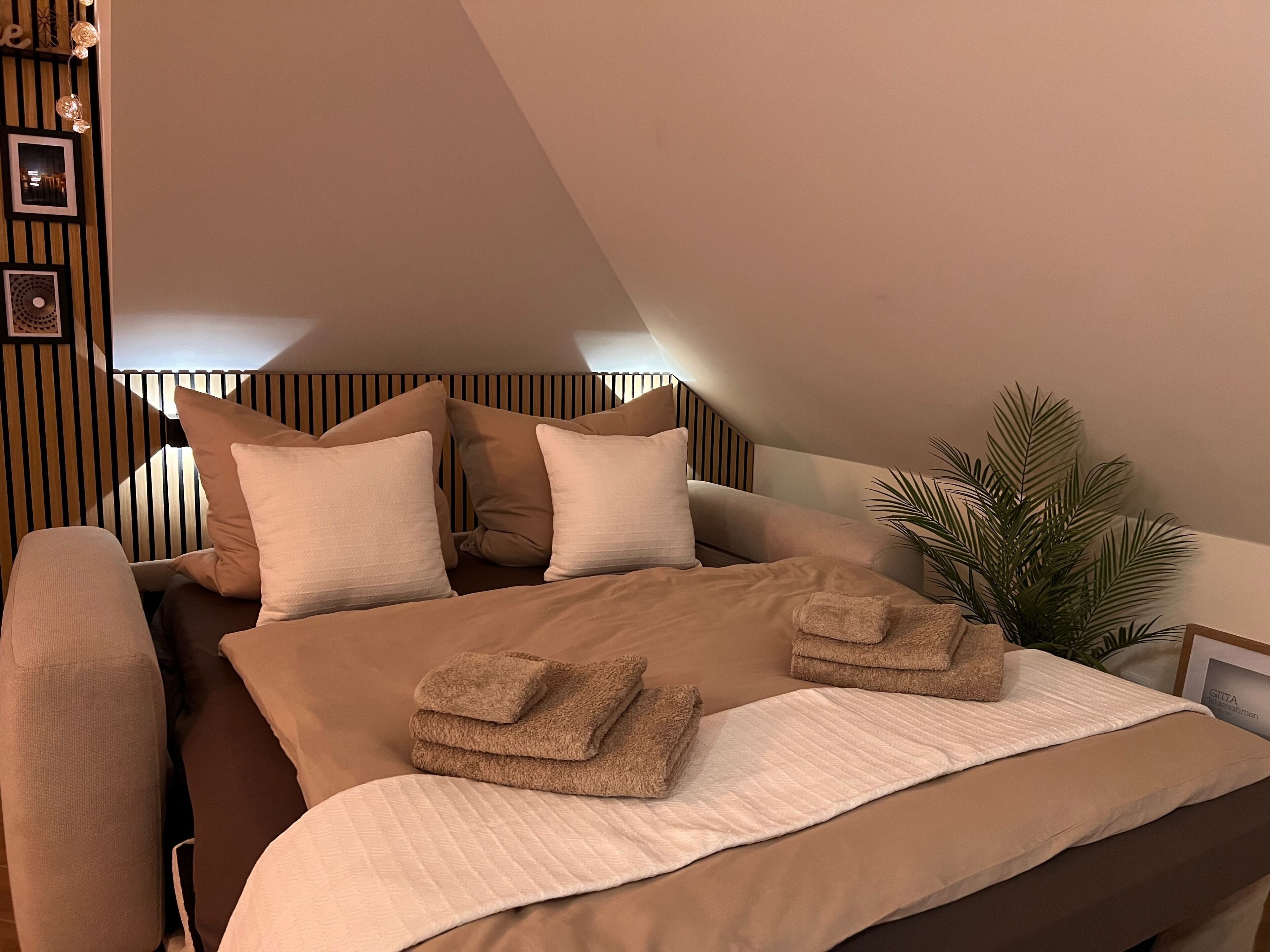 A bedroom with a bed, pillows, a bedside plant, and a wall with vertical lines and framed pictures.