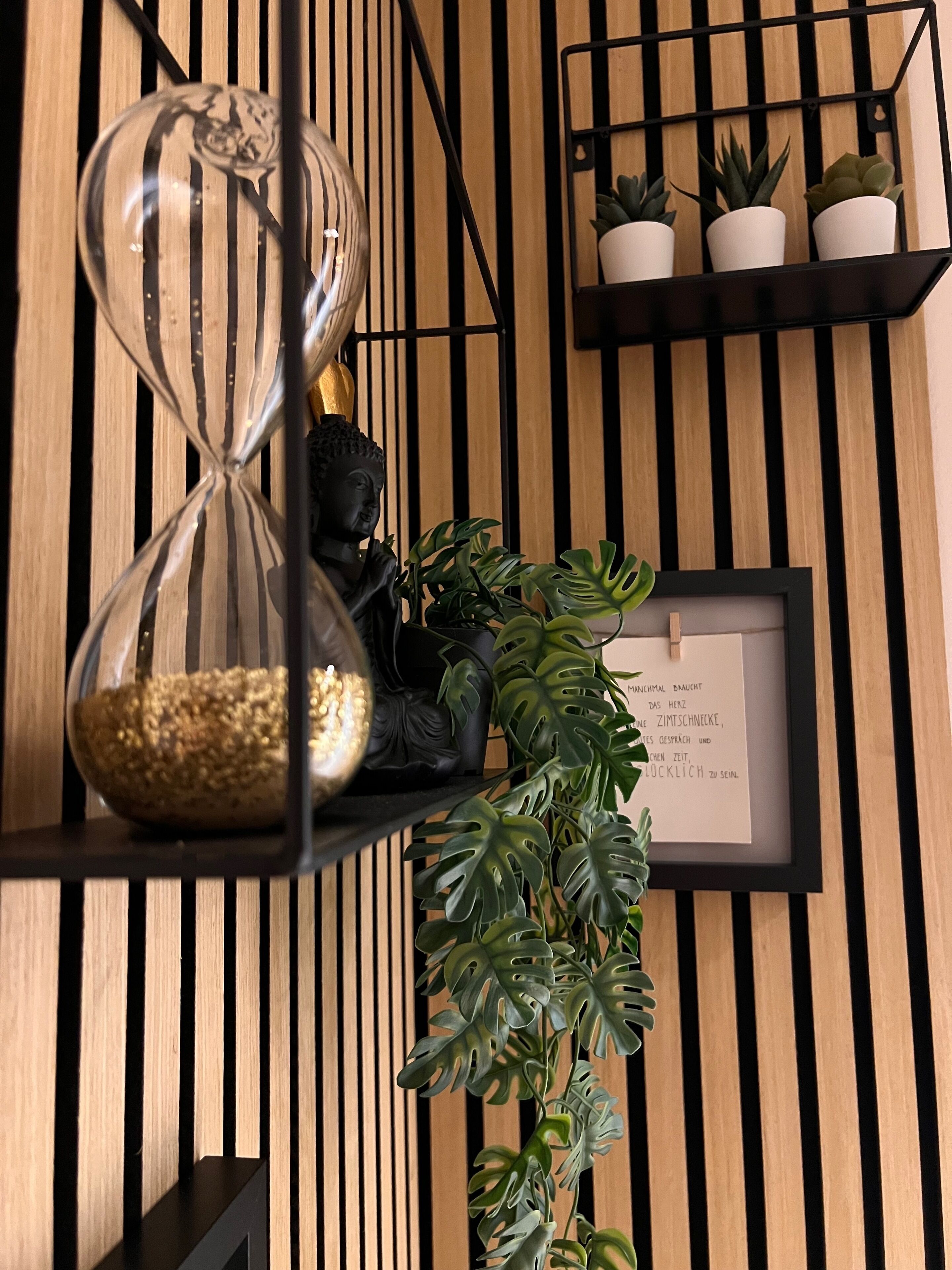 Una pared de madera con listones verticales, una estantería negra con plantas en macetas, una cita enmarcada y una estatua de Buda.