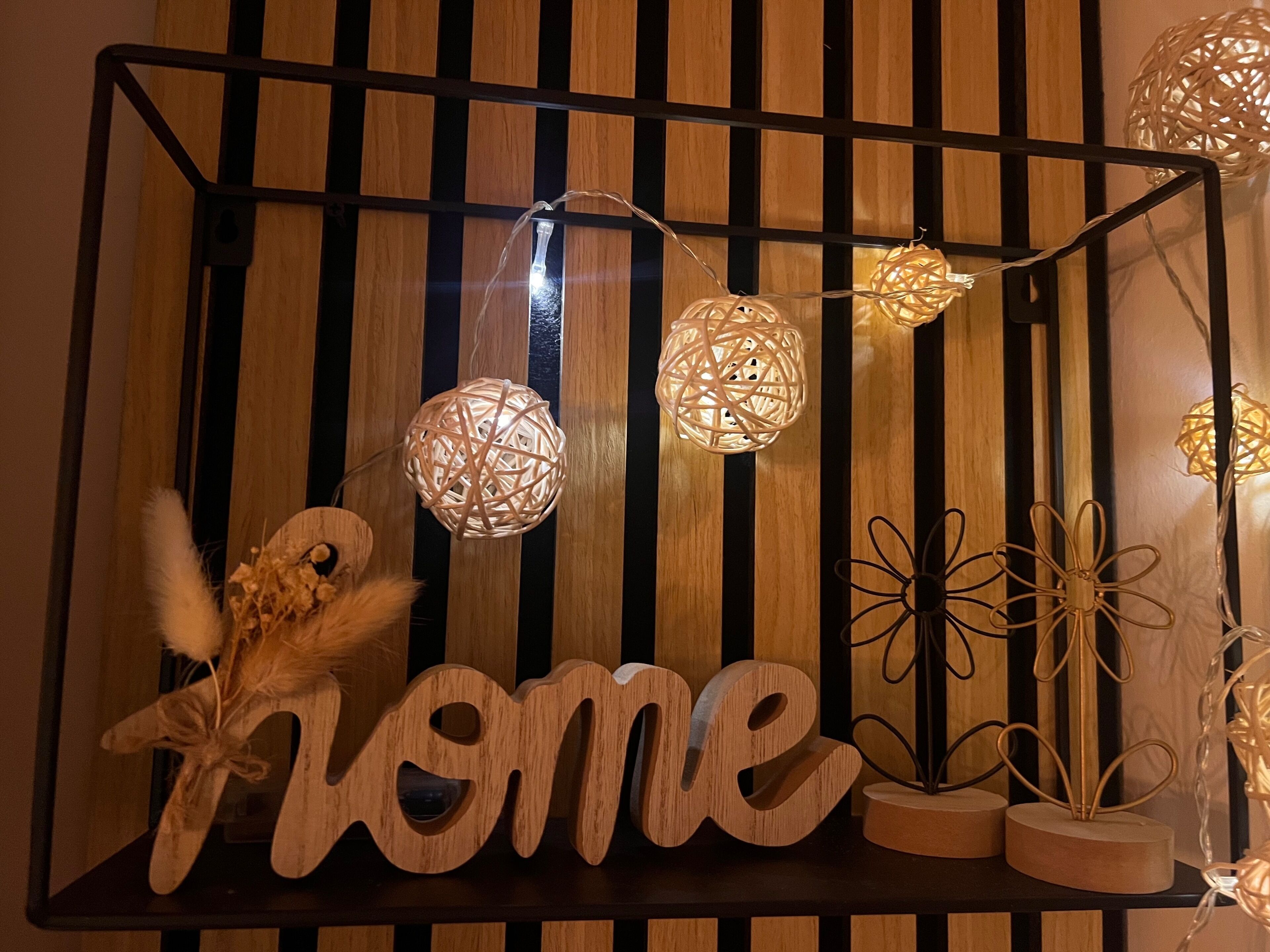 Un mur en bois avec des lattes verticales, des lampes sphériques décoratives et une enseigne en bois portant le mot « chez soi ».