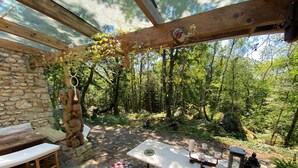 Un área de estar al aire libre rústica, con paredes de piedra, un banco de madera, una mesa con velas y vista a un bosque.