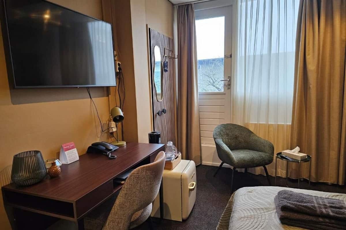 Habitación de hotel con cama, escritorio con silla, televisor, maleta y ventana con cortinas.