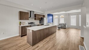 A modern kitchen with a central island, built-in cabinets, and a grey sofa in the adjacent living area. - Brand New 4-BR Ensuite Townhouse | Netflix/Disney+ (Cambridge)