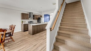 A modern kitchen with wooden cabinets, a central island, and a dining area with a wooden table and chairs. - Brand New 4-BR Ensuite Townhouse | Netflix/Disney+ (Cambridge)