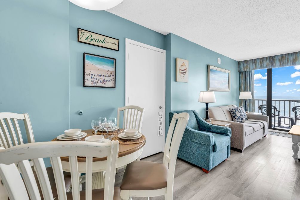 Une pièce comprenant une table à manger dressée pour quatre personnes, un canapé, un fauteuil et une vue sur le ciel grâce à de grandes fenêtres.