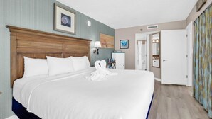 Une chambre d’hôtel avec un grand lit, une tête de lit en bois, une lampe fixée au mur, un tableau encadré et une porte donnant sur une autre pièce.
