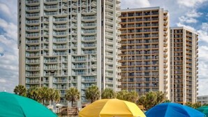 Un hôtel situé en bord de mer, composé de plusieurs immeubles de grande hauteur, avec des chaises de plage, des parasols et des palmiers.