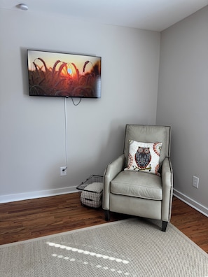 Ein Wohnzimmer mit einem flachbildfernseher an der Wand, einem Ledersessel mit gemusterten Kissen, einem kleinen gewebten Korb und einem grauen Teppich.