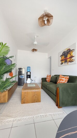 A living room with a green sofa, a wooden coffee table, a refrigerator, a water dispenser, and a ceiling fan. - Olive wood apartment - Mombasa (Mombasa)