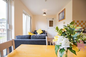 Ein Essbereich mit einem Holztisch, einer Vase mit Blumen, einem grauen Sofa mit gelben Kissen und einer karierten Wand im Hintergrund.