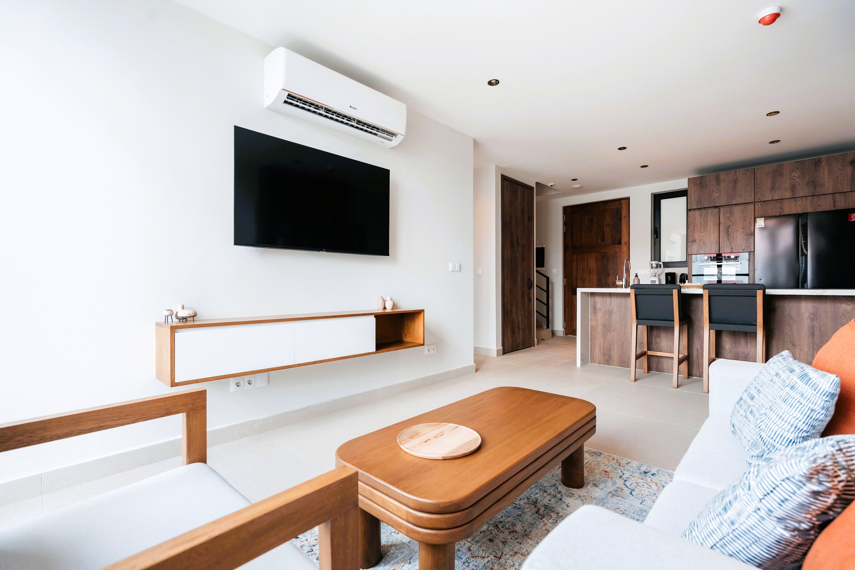 A modern living room with a flat-screen TV, wooden furniture, and a kitchen in the background.