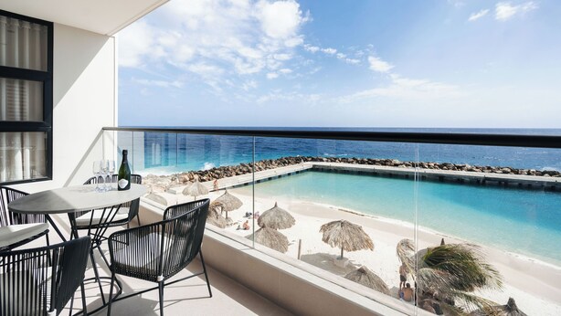 A balcony with a pool, beach view, and outdoor seating. - Bocobay Majestic Apartments (Willemstad)