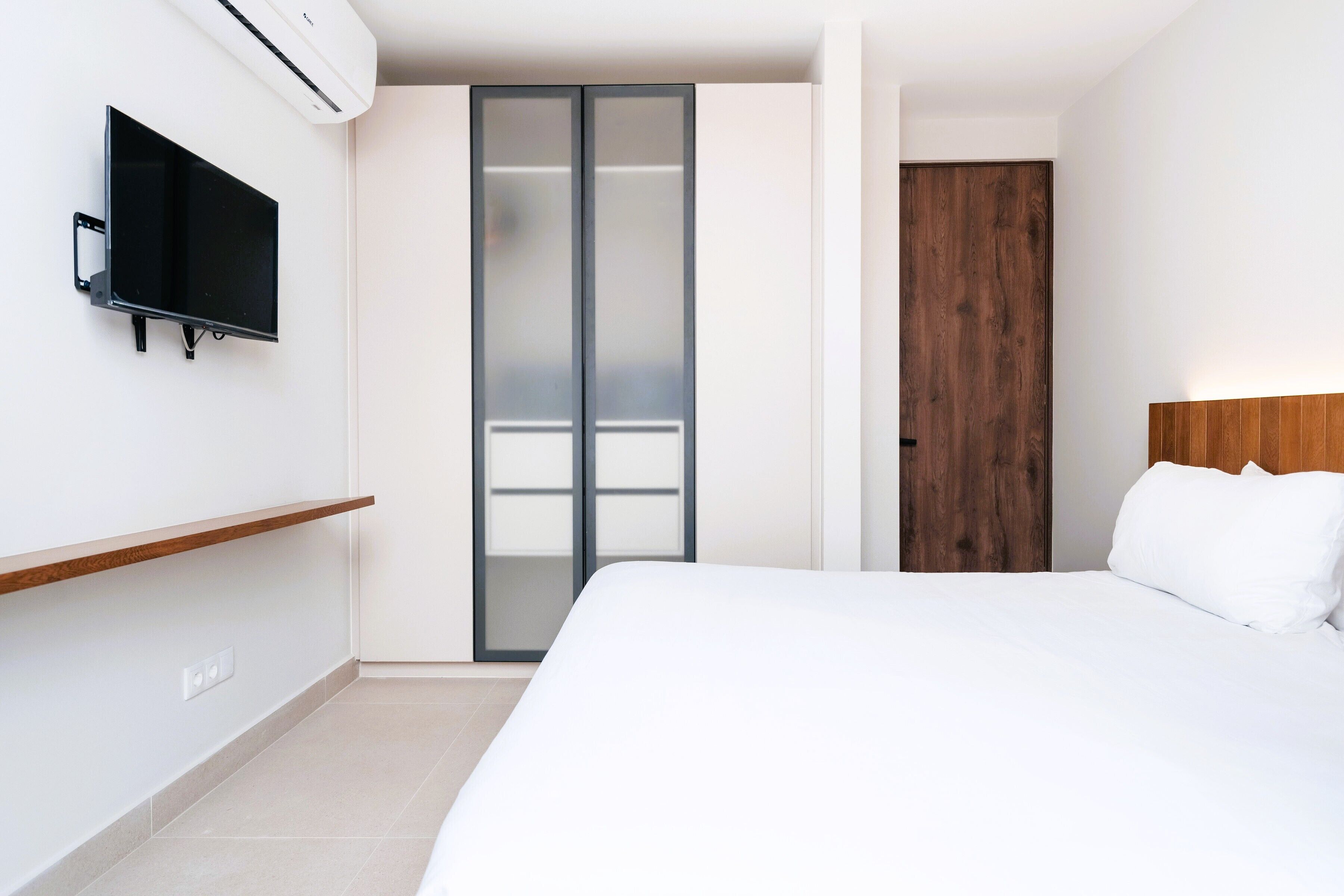 A modern hotel room with a bed, a TV mounted on the wall, a sliding door, and a wooden headboard.