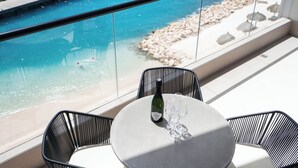 A balcony with a glass railing overlooking a pool and beach, a table with a bottle and glasses, and black chairs. - Bocobay Majestic Apartments (Willemstad)