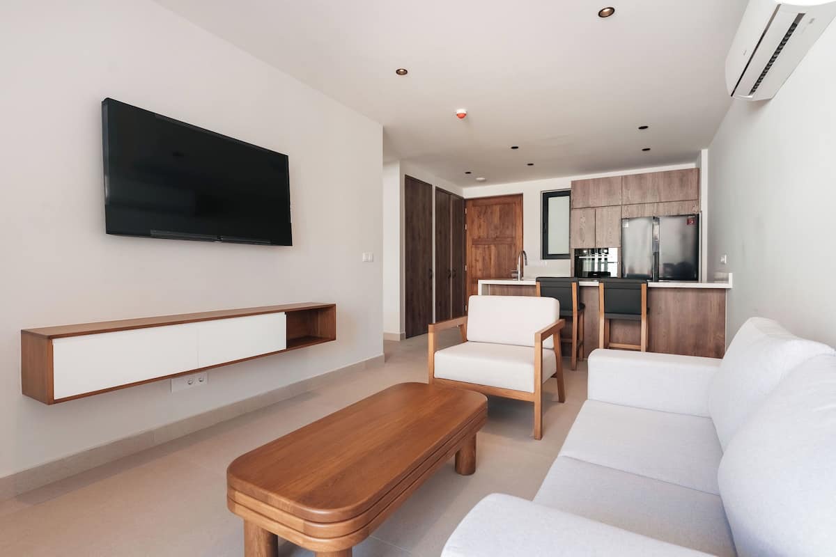 Un salon moderne avec un téléviseur à écran plat, une table basse en bois, un canapé blanc et un coin cuisine à l’arrière-plan.