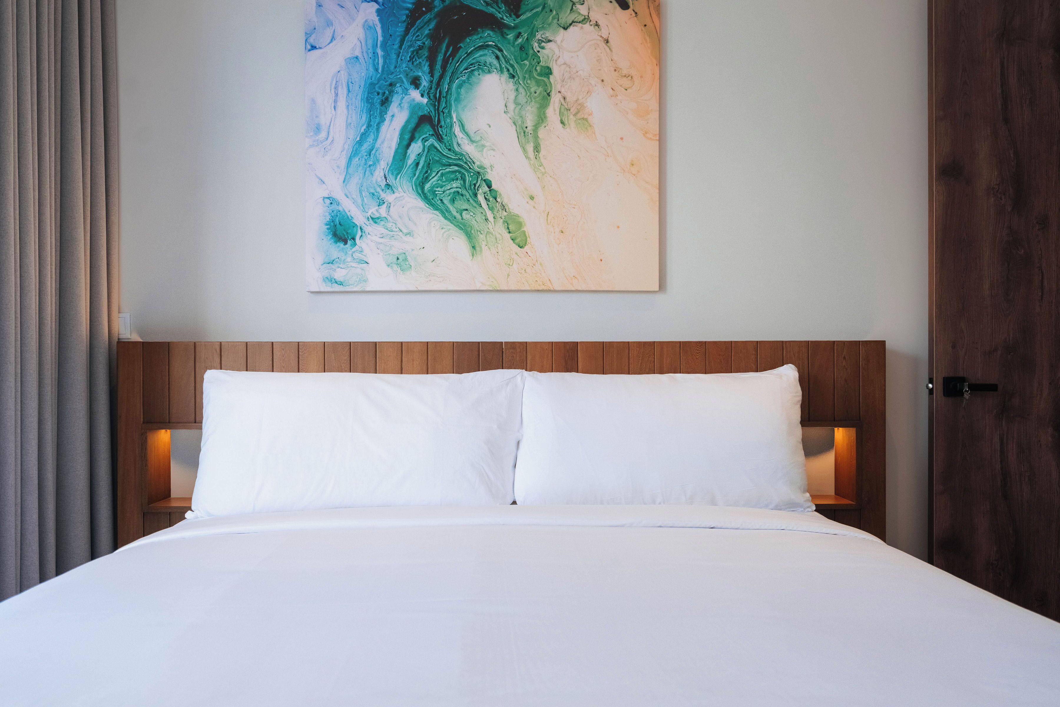 A bed with white linens, a wooden headboard, and a wall-mounted abstract painting.
