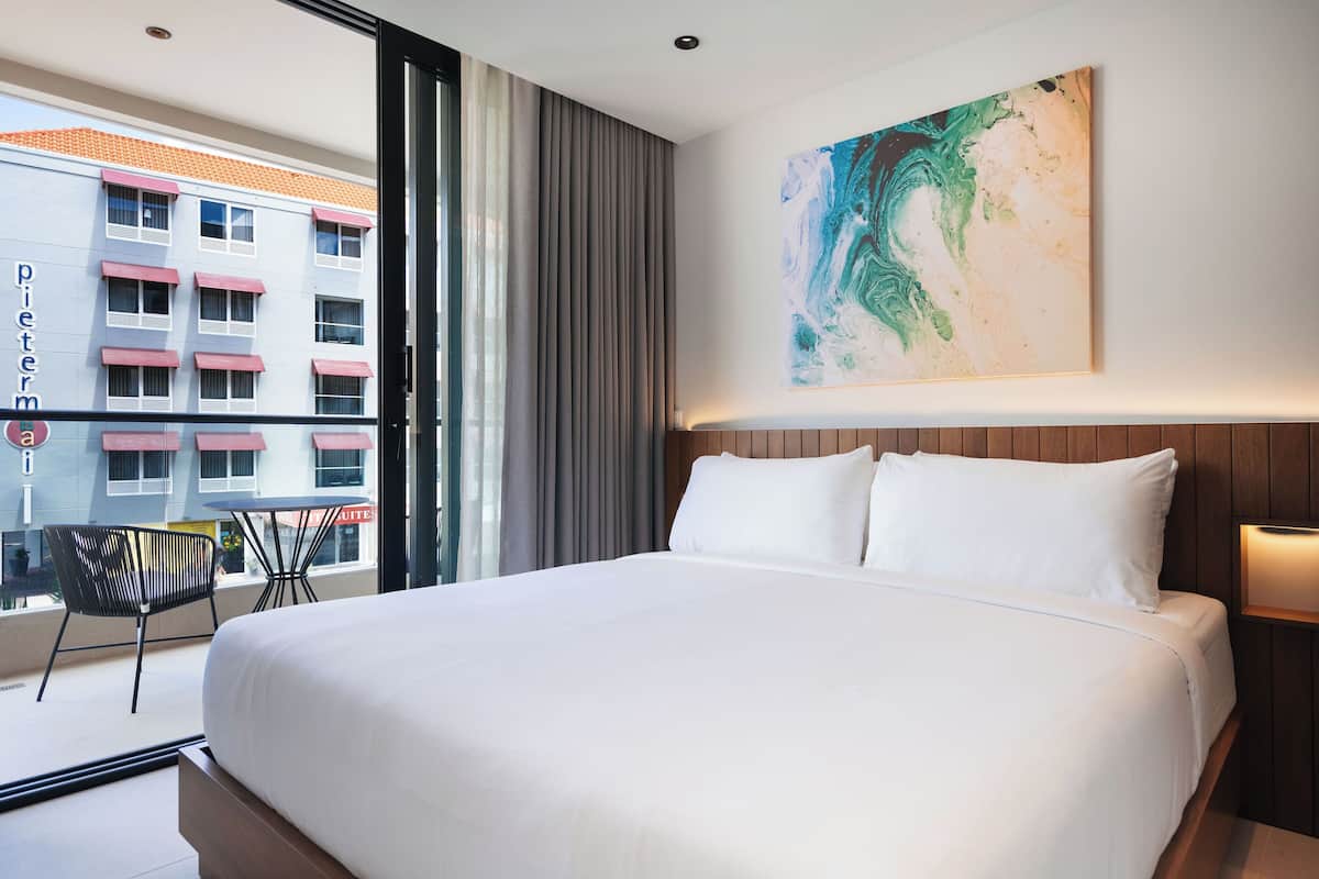 Une chambre d’hôtel avec un grand lit, une vue sur un immeuble doté de balcons, une peinture abstraite moderne et une chaise près de la fenêtre.