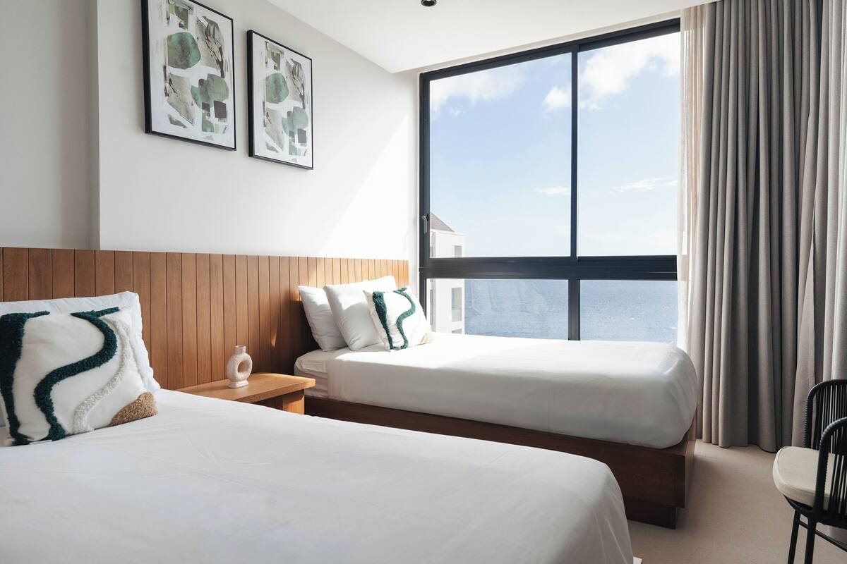 Une chambre d’hôtel avec deux lits, une tête de lit en bois, une table de chevet, une chaise et une grande fenêtre avec des rideaux.
