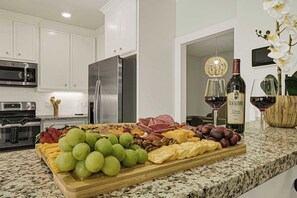 Uma cozinha com armários brancos, eletrodomésticos de aço inoxidável, bancada de mármore e uma tábua de frutas e queijos.