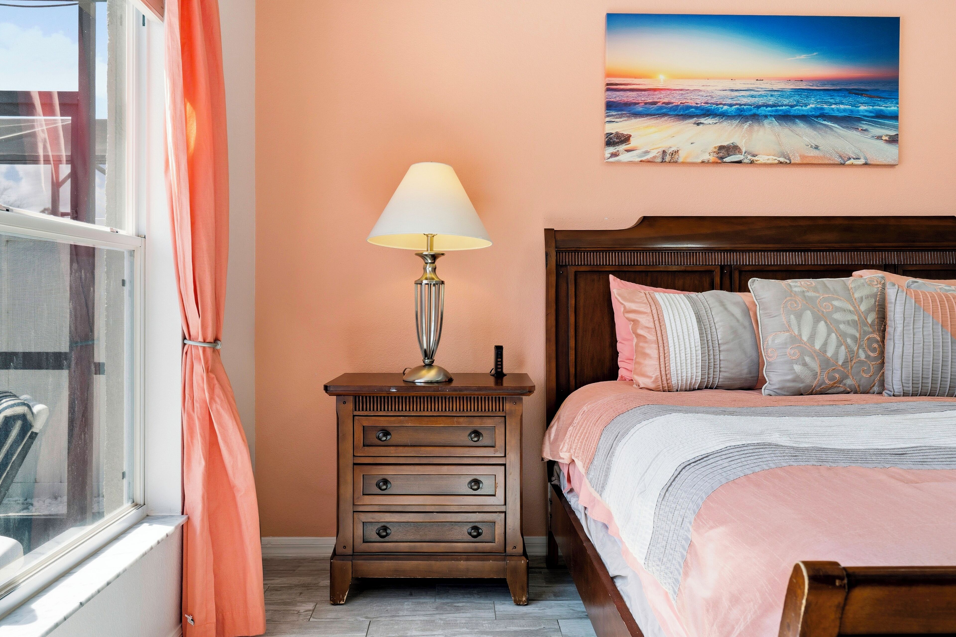 A bedroom with a bed, a nightstand with a lamp, a window with curtains, and a painting on the wall.
