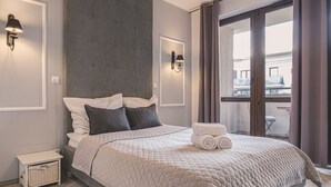Une chambre moderne avec un grand lit, une tête de lit, deux lampes fixées au mur, une petite table de chevet et une vue sur l’extérieur grâce à une porte-fenêtre coulissante.
