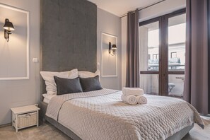 Une chambre moderne avec un grand lit, une tête de lit, deux lampes fixées au mur, une petite table de chevet et une vue sur l’extérieur grâce à une porte-fenêtre coulissante.