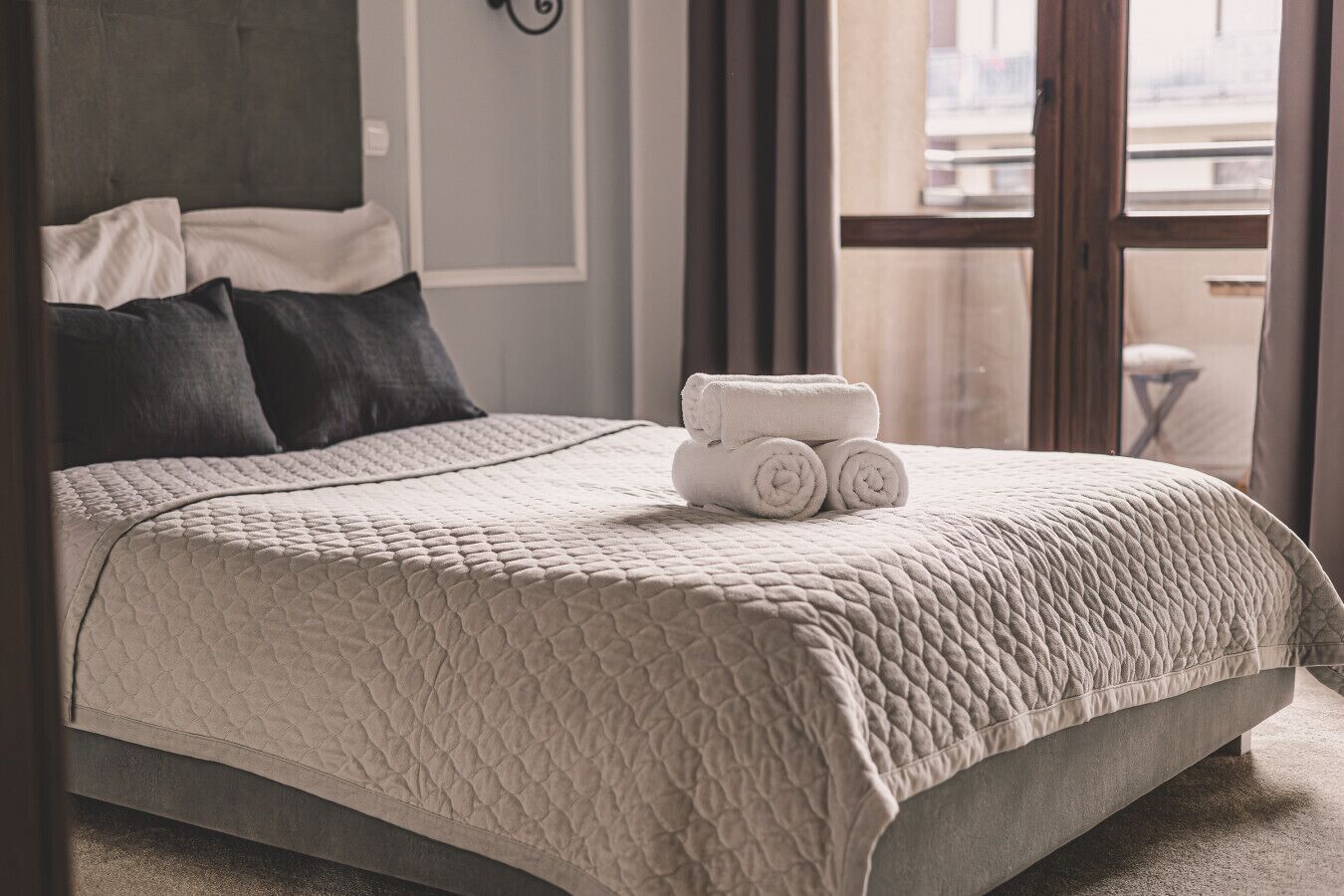 A neatly made bed with a quilted cover, a stack of rolled towels, and a view of the outside through a window.