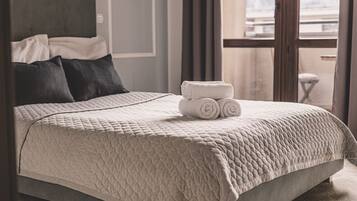 A neatly made bed with a quilted cover, a stack of rolled towels, and a view of the outside through a window.