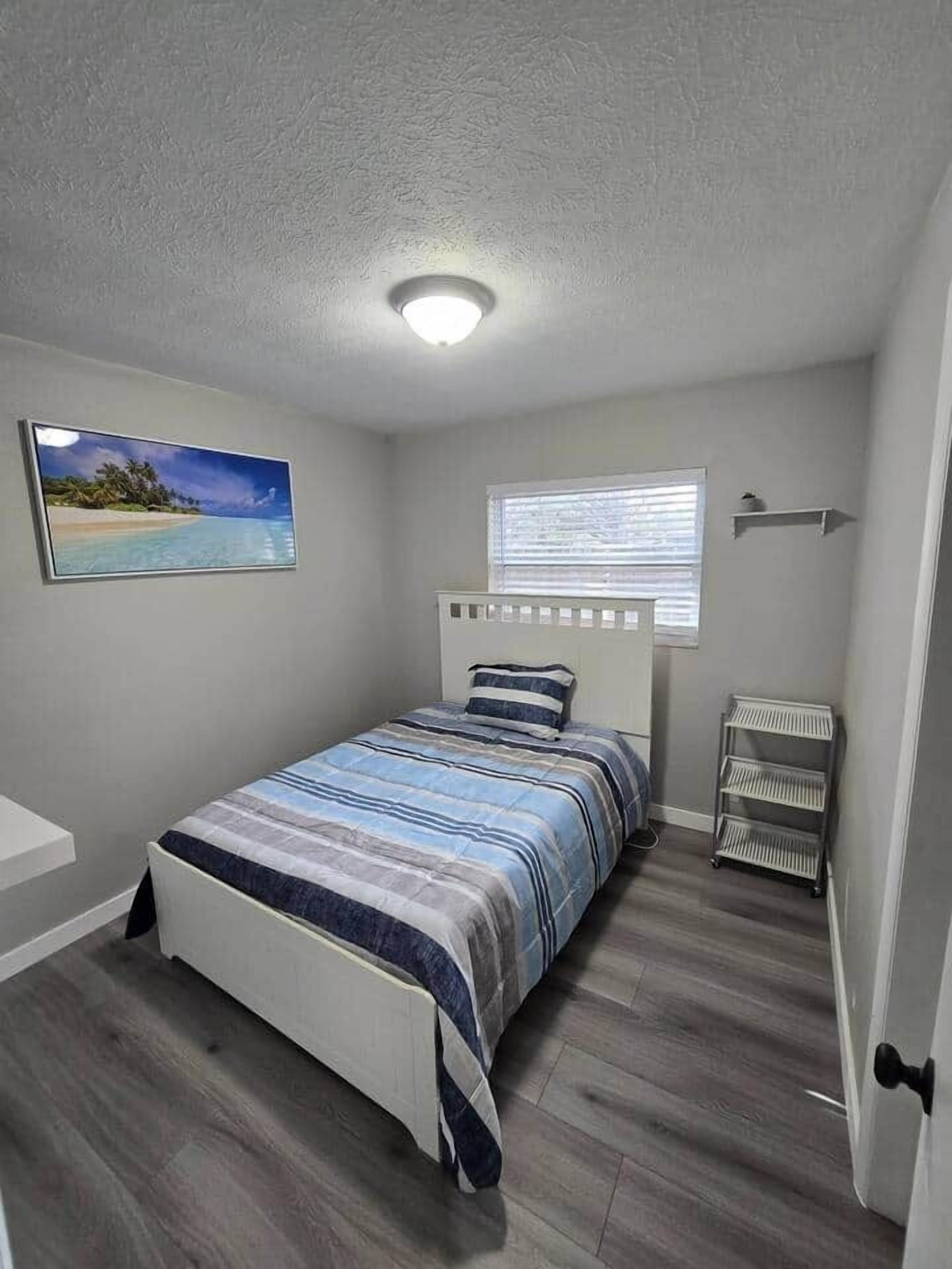 A bedroom with a bed, a window with blinds, a wall-mounted shelf, and a painting of a beach scene.