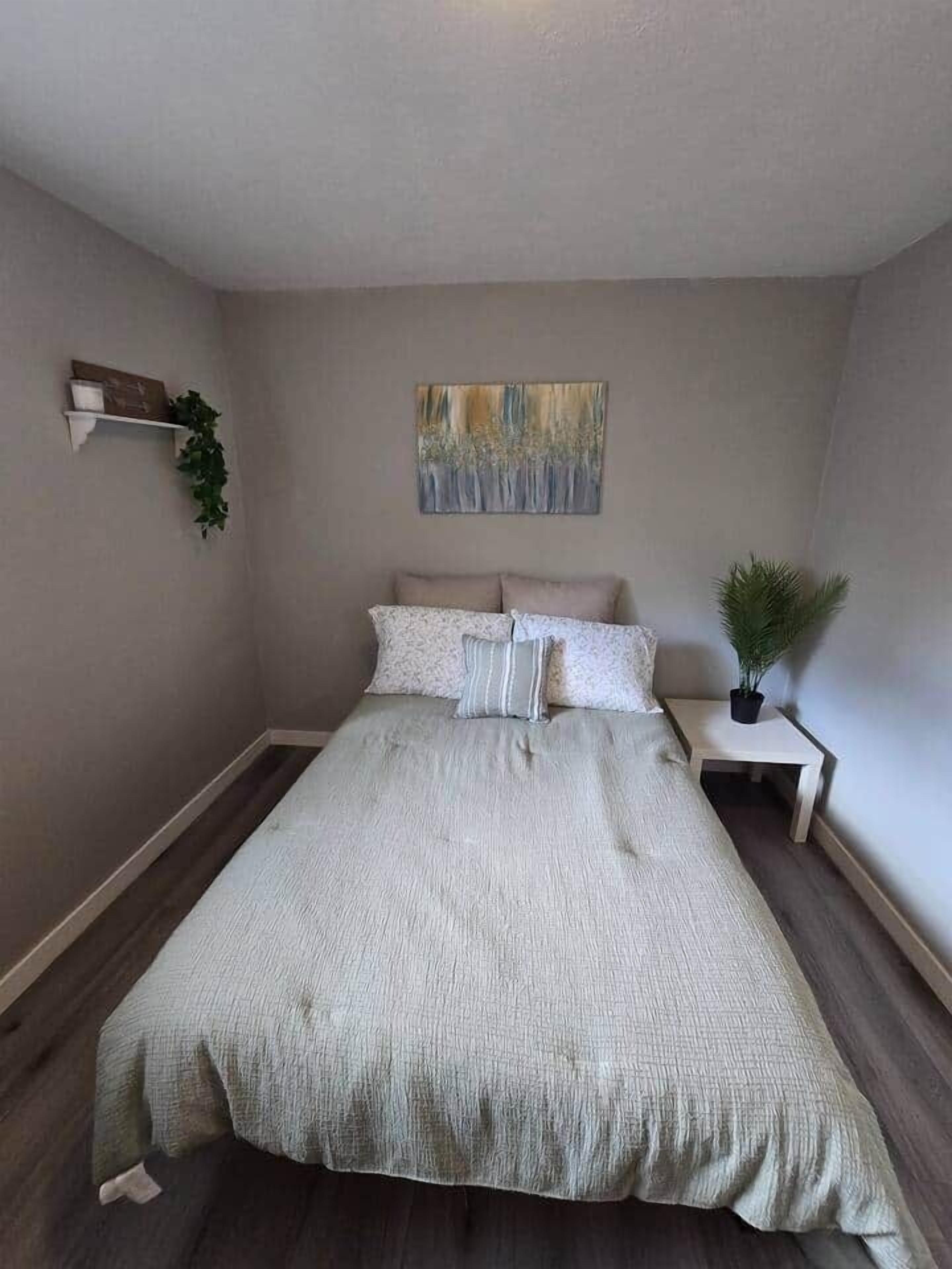 A bedroom with a bed, a nightstand, a wall-mounted shelf with a plant, and a painting.