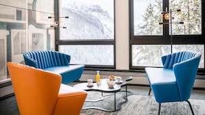 Ett modernt rum med stora fönster, två blå fåtöljer, en orange stol, ett soffbord och ett element.