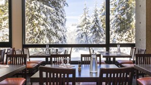 En restaurang med bord och stolar i trä, och en fri utsikt över snötäckta träd genom stora fönster.
