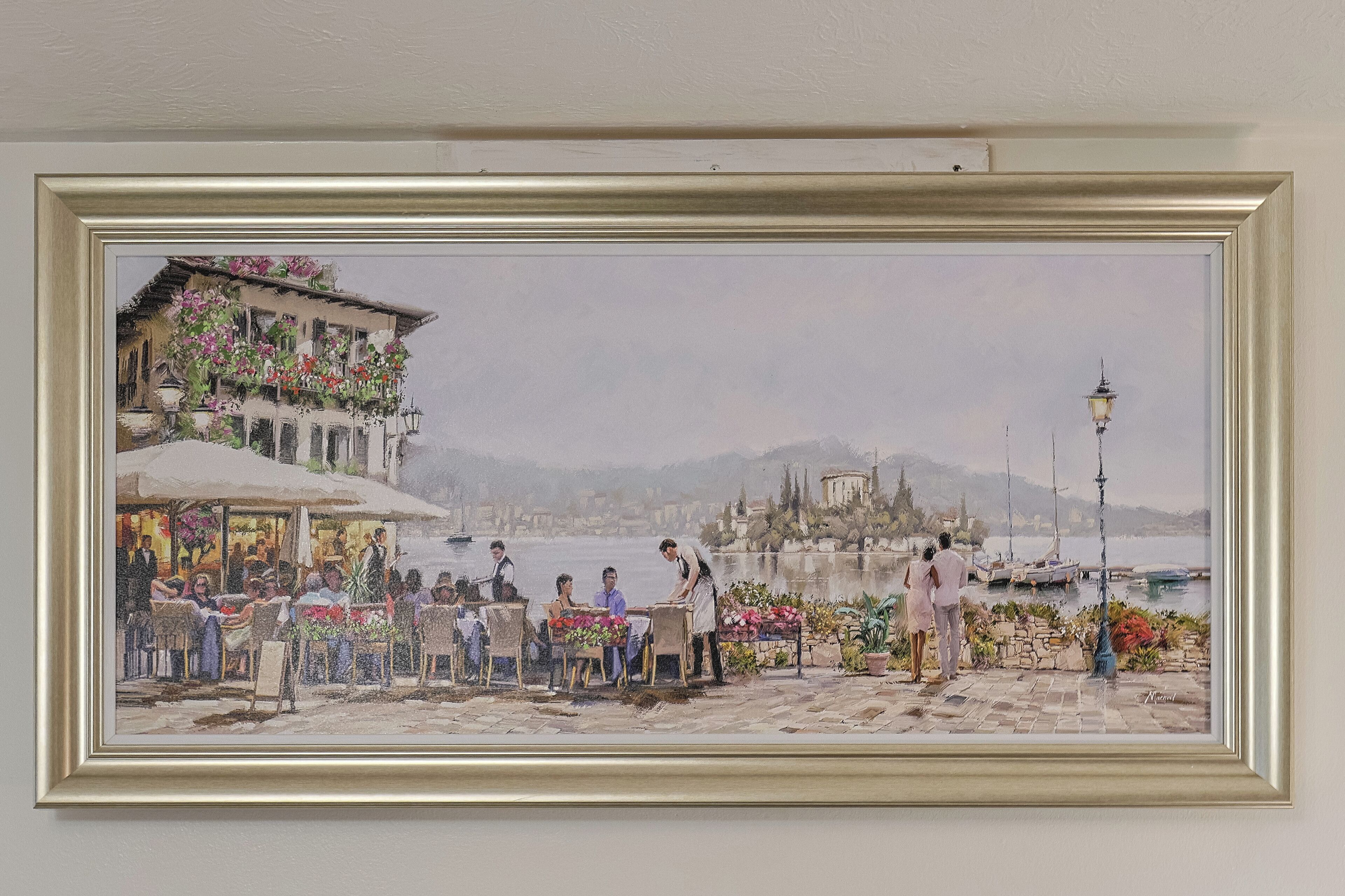 A framed painting of a coastal scene with people, a building with flowers, and a body of water.