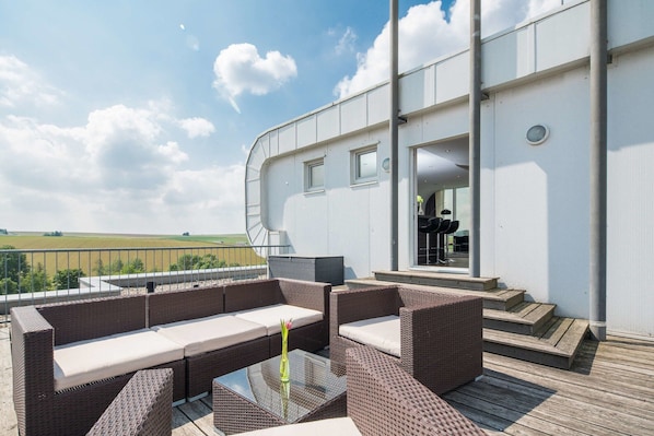 Eine Terrasse im Freien mit Korbmöbeln und einem Glastisch, mit Blick auf ein Feld.