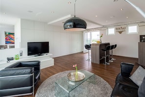 A modern living room with a TV, black leather sofas, a glass coffee table with a vase, and a kitchen area with bar stools. - Hotel Bad Rappenau (Bad Rappenau)