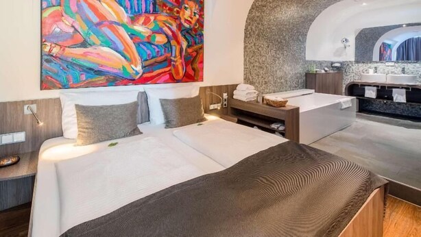 A modern hotel room with a large bed, a small desk, a bathroom with a vanity, and a colorful abstract painting on the wall. - Hotel Bad Rappenau (Bad Rappenau)
