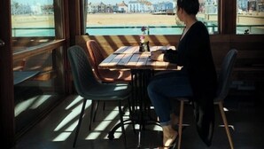 Eine Person sitzt an einem Tisch mit Blick auf einen Strand und Gebäude durch große Fenster.
