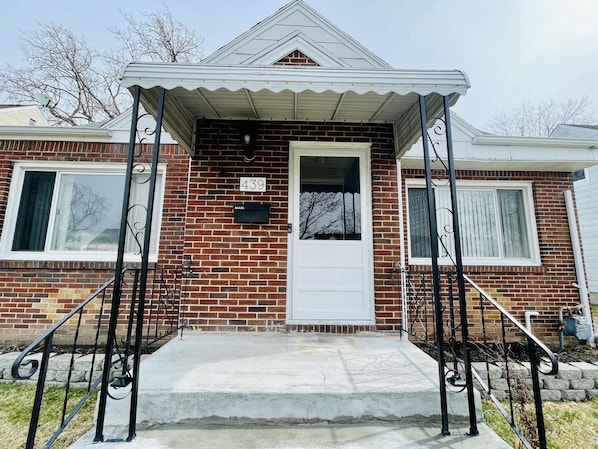 A two-story brick house with a white front door, a mailbox, and a covered porch with metal railings. - Comfortable 3BR House Near UB, Parking, WiFi, Backyard (Buffalo)