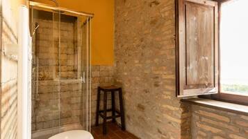 Un baño con ducha, inodoro, un taburete de madera y una ventana.