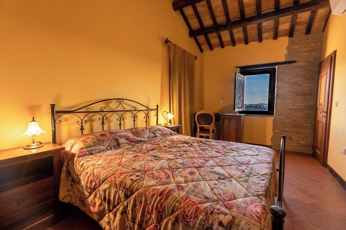 A bedroom with a bed, a nightstand, a lamp, a chair, a desk, and a window with a view.