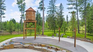Eine hölzerne Wasserturmkonstruktion in einem Park mit gepflastertem Bereich, Sitzgelegenheiten und einem Kiesweg.