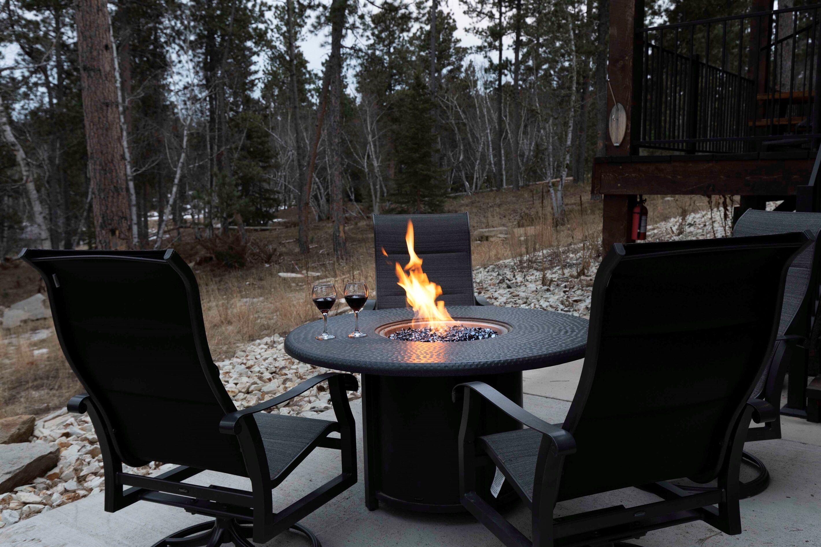 A round outdoor table with a built-in fire feature, surrounded by two black chairs.